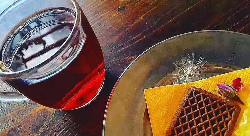 نوشیدنی گرم، وافل شکلاتی، و غنچه گل رز روی میز چوبی کافه رد. لحظات شیرین و آرامش‌بخش در فضایی دلنشین.