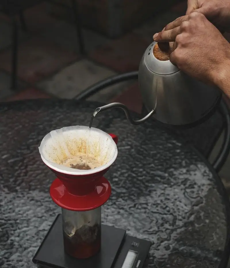 تصویری زیبا از دم آوری دقیق قهوه دمی توسط باریستا در کافه رد. آماده سازی قهوه تخصصی با V60 و کتری گردن غازی روی ترازو.