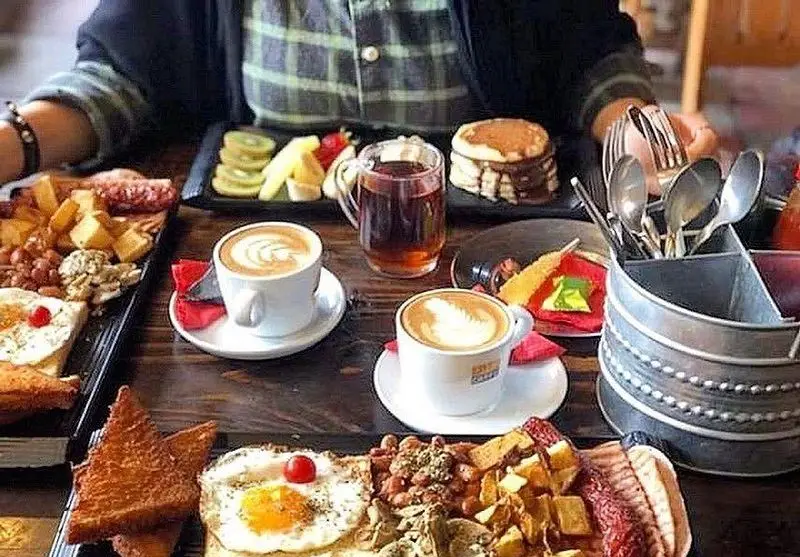 یک میز صبحانه کامل و رنگارنگ در کافه رد: دو فنجان قهوه لاته، پنکیک، بشقاب میوه، تخم مرغ سرخ شده، لوبیا و نان تست.