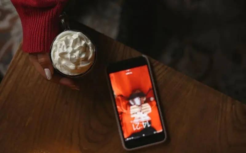 تجربه ای دلنشین در کافه رد: لیوان نوشیدنی خامه دار و گوشی موبایل روی میز چوبی. لذت بردن از لحظات آرام در فضای دنج و گرم کافه.