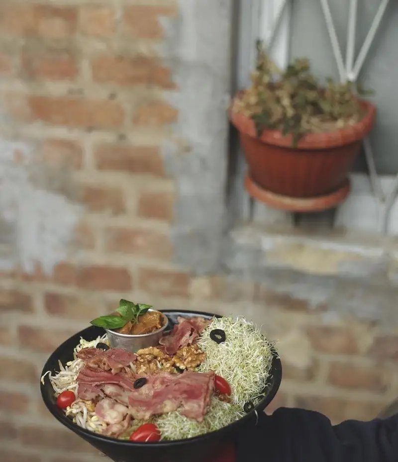 تصویری جذاب از سالاد ویژه کافه رد شامل ژامبون، گردو، جوانه و سبزیجات تازه در محیطی دلنشین کنار دیوار آجری.