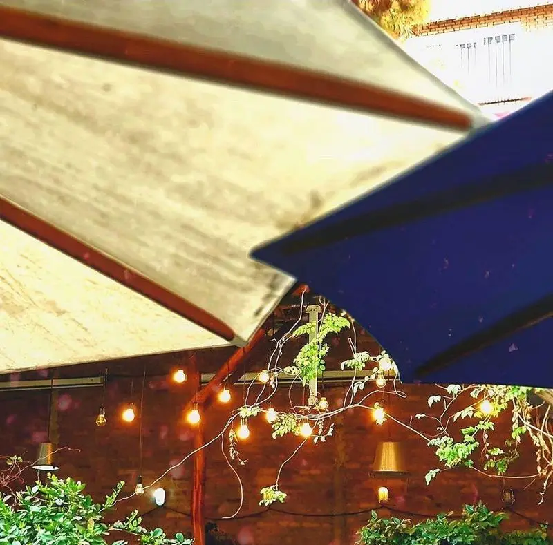 فضای باز و دنج کافه رد با چترهای بزرگ کرم و آبی، نورپردازی گرم با ریسه لامپ و گیاهان سبز. محیطی ایده آل برای کافه گردی در کافه رد.