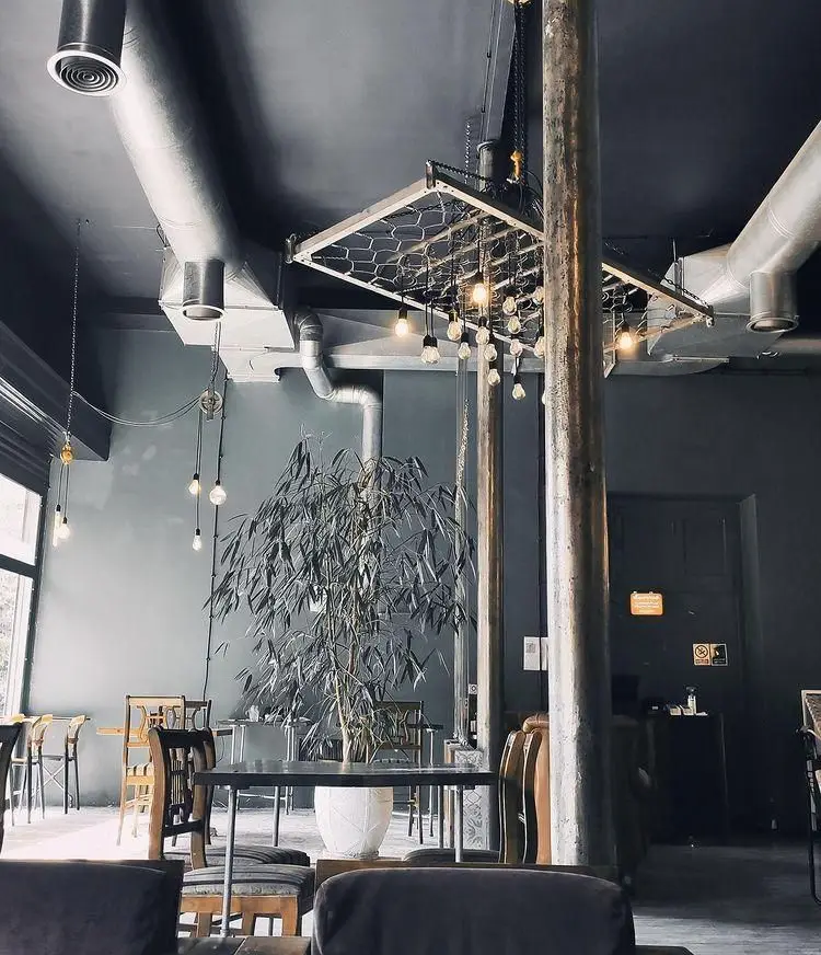 نمای داخلی کافه رد با دکوراسیون صنعتی و مدرن، سقف سیاه، کانال‌های تهویه آشکار و نورپردازی خاص با لامپ‌های آویز، همراه با گیاه بزرگ و فضای آرامش‌بخش.