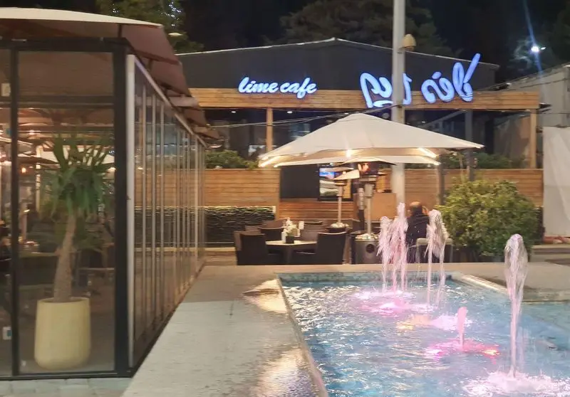 نمای جذاب شب از کافه رستوران لایم، شامل فضای بیرونی دنج با میز و صندلی، چتر، فواره رنگی و نورپردازی زیبا.
