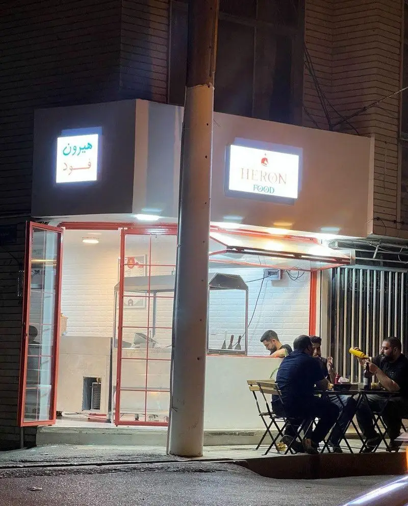 فست فود هیرون فود در شب، با تابلوهای روشن فارسی و انگلیسی. مشتریان در حال لذت بردن از پیتزا و ساندویچ در فضای باز.