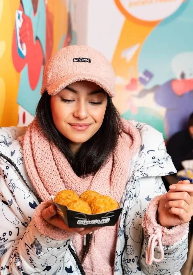 زن جوانی با کلاه صورتی در دیب چیز شعبه سعادت آباد، با لبخندی دلنشین به توپک‌های پنیری طلایی و خوشمزه خود می‌نگرد. لذت غذایی خاص.