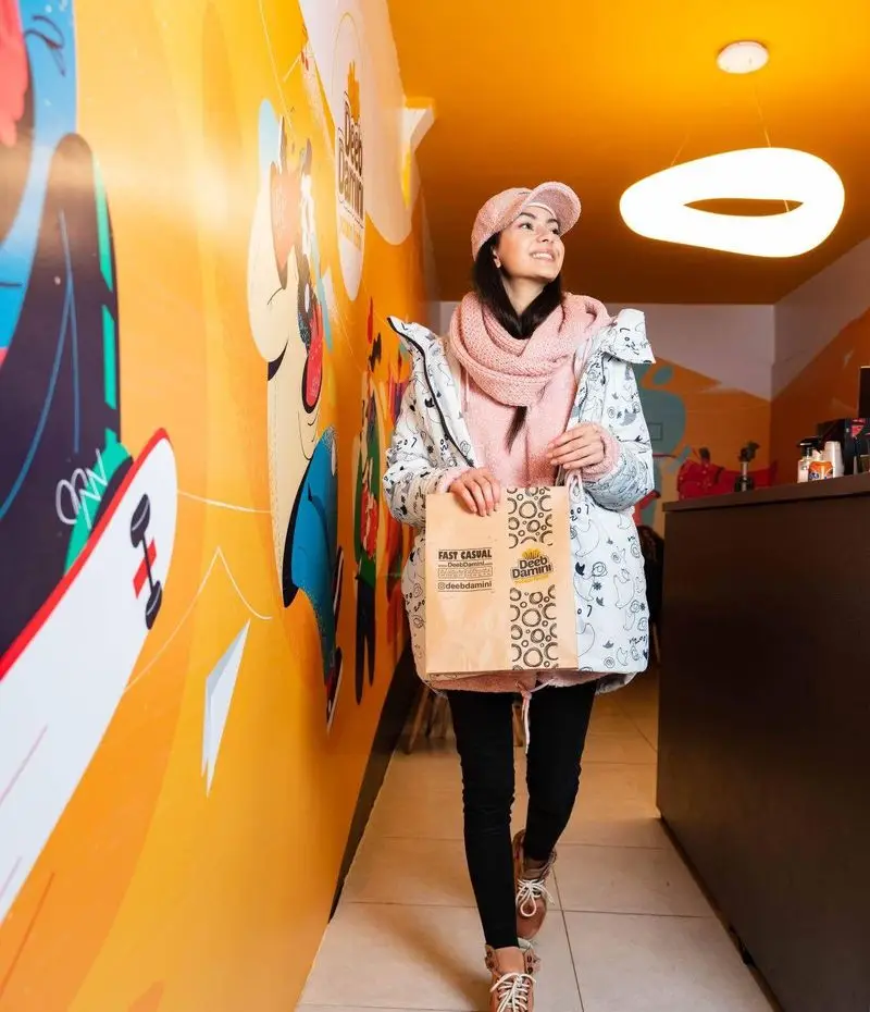 زنی خوش‌پوش با کیسه دیب چیز (Deeb Damini) در فضای شاد و مدرن شعبه سعادت آباد، با دیوارنگاری‌های رنگارنگ و سقف نارنجی.