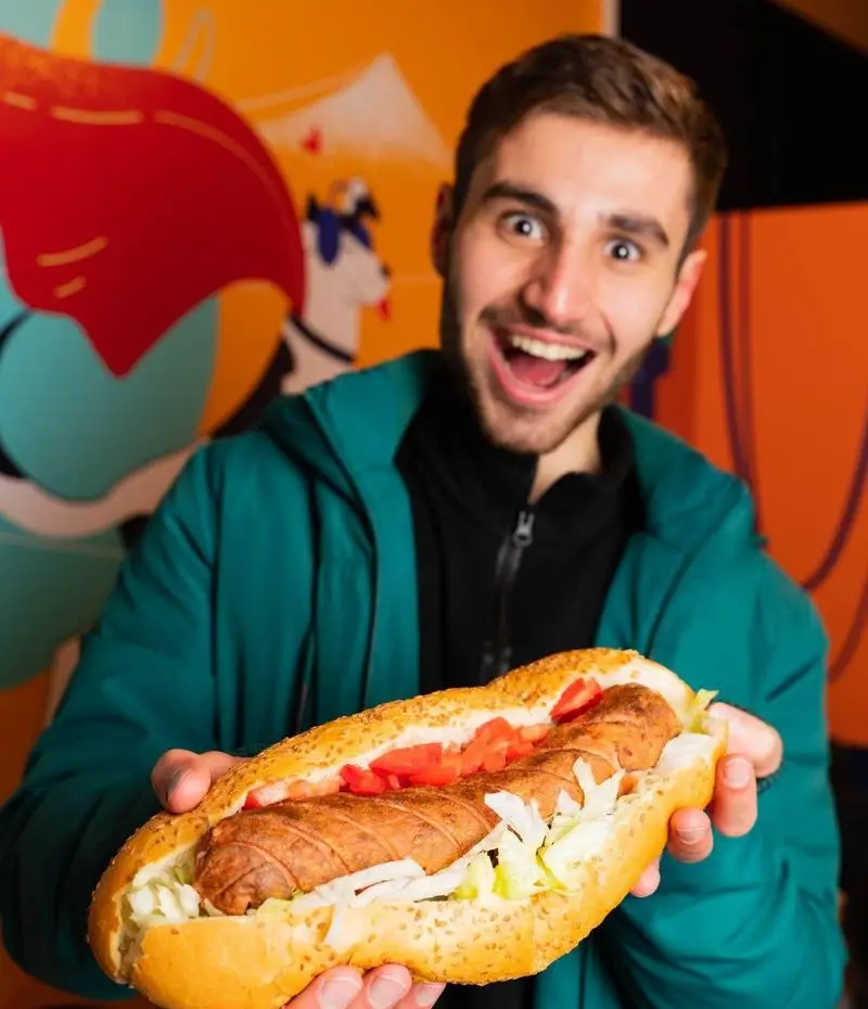 مشتری خوشحال دیب چیز سعادت آباد ساندویچ سوسیس بزرگ و تازه را با کاهو و گوجه فرنگی با هیجان نمایش می‌دهد.