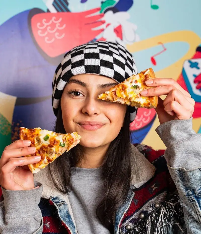 دختر جوانی با کلاه شطرنجی در دیب چیز شعبه سعادت آباد لبخند می‌زند و دو اسلایس پیتزای خوشمزه در دست دارد که آماده خوردن هستند.