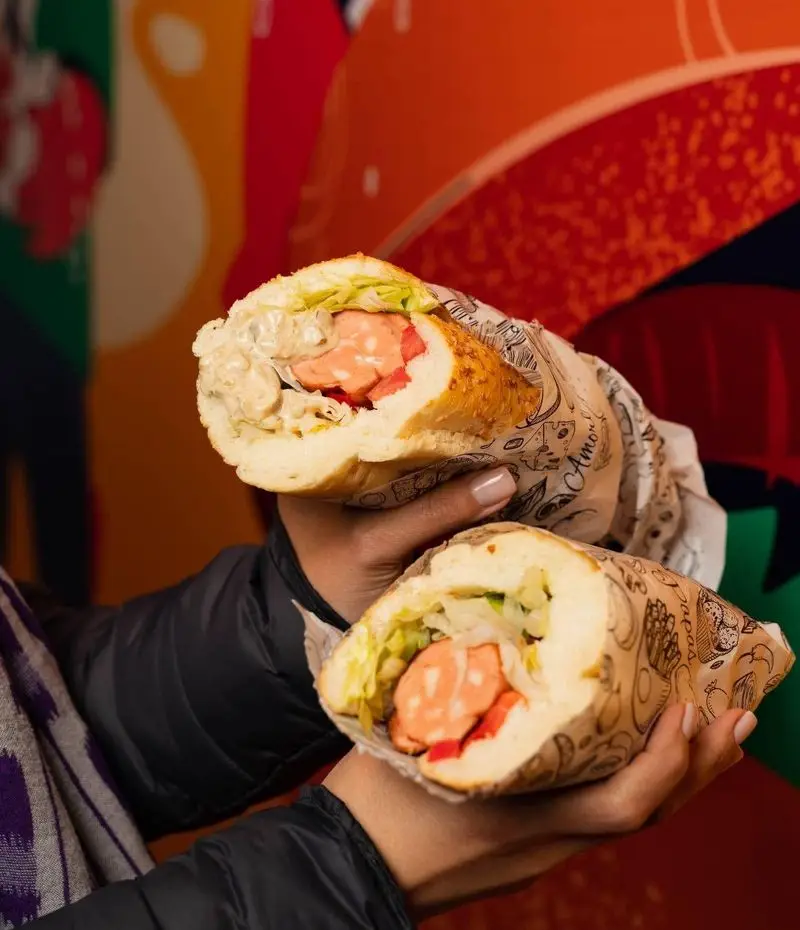 دو ساندویچ بزرگ و خوشمزه از دیب چیز شعبه سعادت آباد، با هات داگ، سبزیجات تازه و سس‌های لذیذ در دست مشتری، آماده سرو.