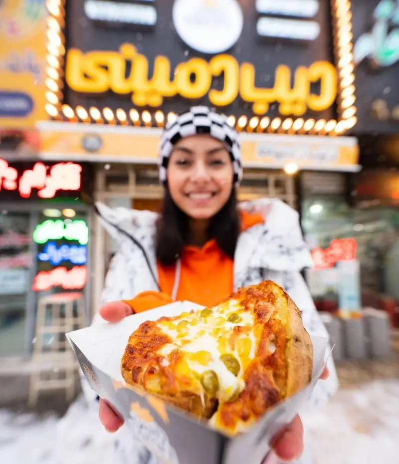 دختر جوان شاد با کلاه شطرنجی، سیب زمینی پنیری داغ دیب چیز شعبه سعادت آباد را در روز برفی با لبخند نشان می دهد.