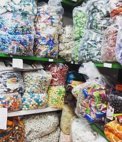 تصویری از قفسه‌های پر از شیرینی‌جات، آبنبات‌ها و خشکبار رنگارنگ در بسته‌بندی‌های شفاف در فروشگاه شیرینی‌جات بخشی.