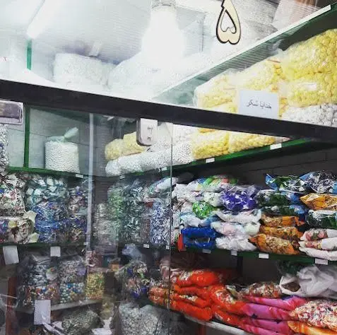 تصویری از قفسه‌های پر و متنوع فروشگاه شیرینی‌جات بخشی، شامل بسته‌های رنگارنگ پشمک، نقل، آجیل و انواع تنقلات خوشمزه.