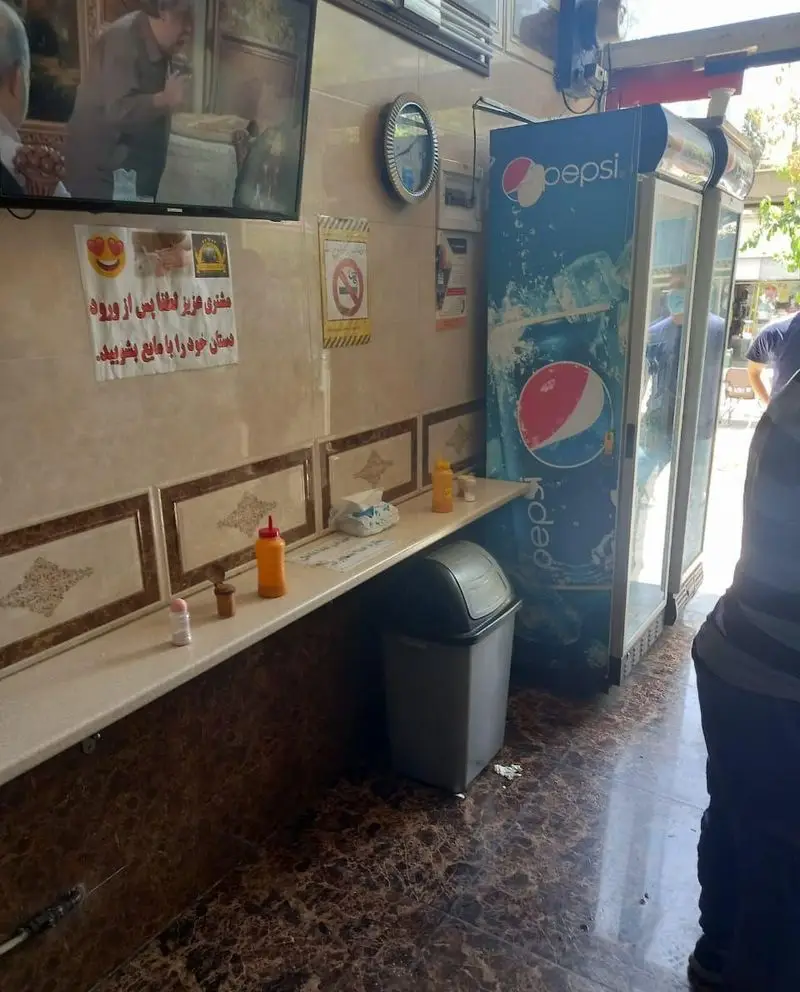 فضای داخلی دنج و تمیز فلافل ابوحیدر با تلویزیون در حال پخش برنامه، یخچال پپسی و پیشخوان مجهز به ضدعفونی‌کننده دست.