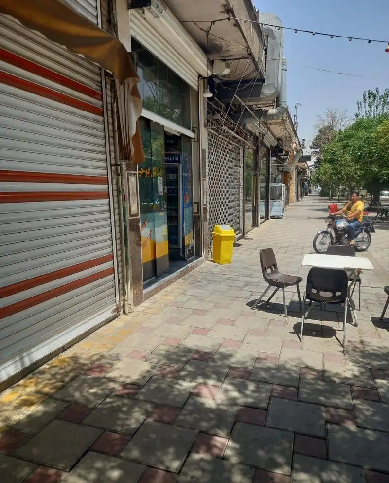 نمای بیرونی مغازه فلافل ابوحیدر در خیابان، با کرکره نیمه باز، یخچال پپسی، و میز و صندلی برای نشستن مشتریان در فضای باز.