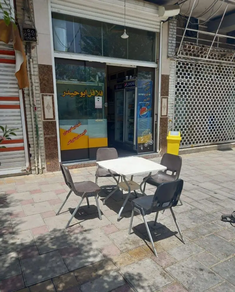 تصویری از نمای بیرونی فلافل ابوحیدر با تابلوی فارسی، میز و صندلی در پیاده‌رو برای پذیرایی مشتریان و یخچال پپسی در ورودی.