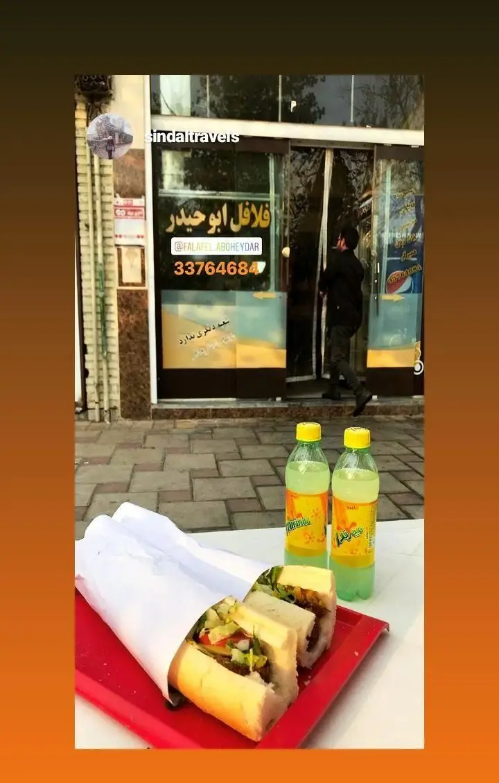 ساندویچ‌های فلافل تازه و خوشمزه فلافل ابوحیدر روی سینی قرمز با نوشیدنی‌های خنک. نمای ورودی فروشگاه در پس‌زمینه.