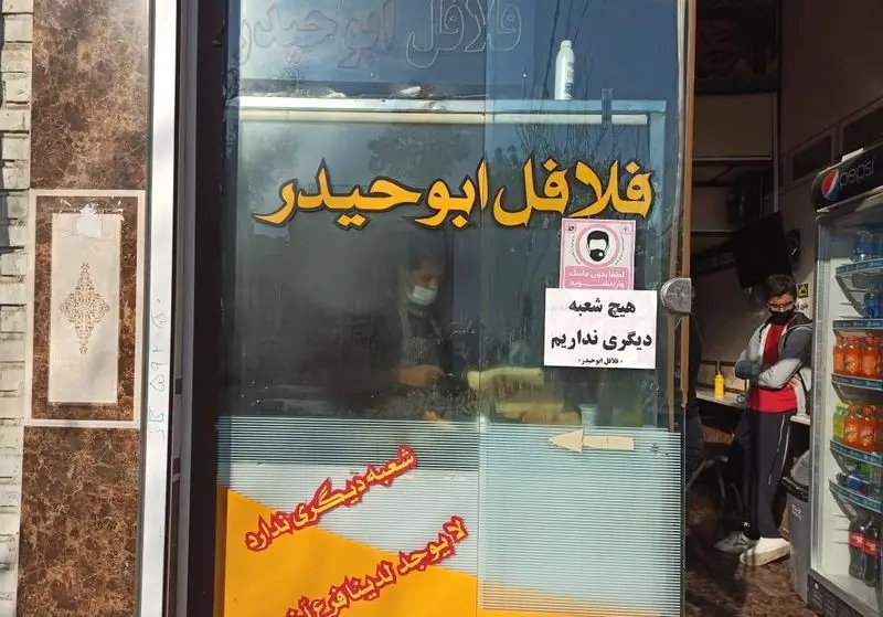 نمای بیرونی مغازه فلافل ابوحیدر با تابلوی زرد رنگ، کارمند ماسک‌زده در حال تهیه غذا و مشتری کنار یخچال نوشیدنی.