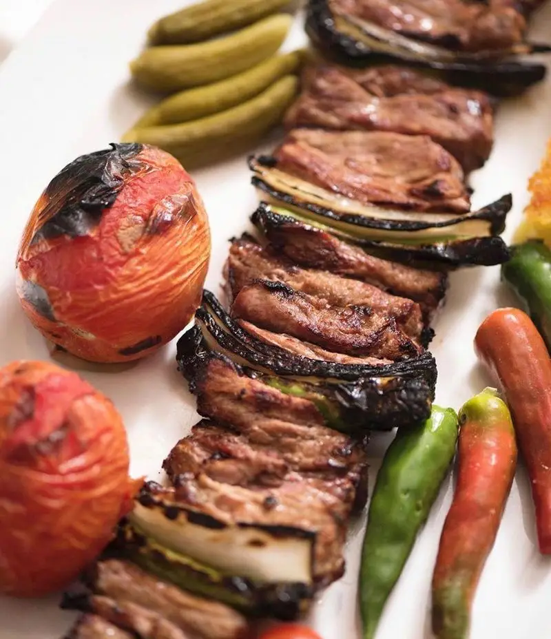 تصویری جذاب از کباب گوشت آبدار به همراه پیاز، گوجه و فلفل کبابی، در کنار خیارشور. تجربه طعم اصیل در رستوران نایب ابان.