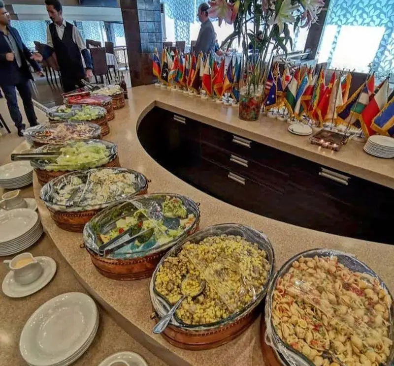میز بوفه سلف سرویس متنوع رستوران نایب ابان شامل انواع سالاد، پاستا و غذاها، در کنار پرچم‌های بین‌المللی.