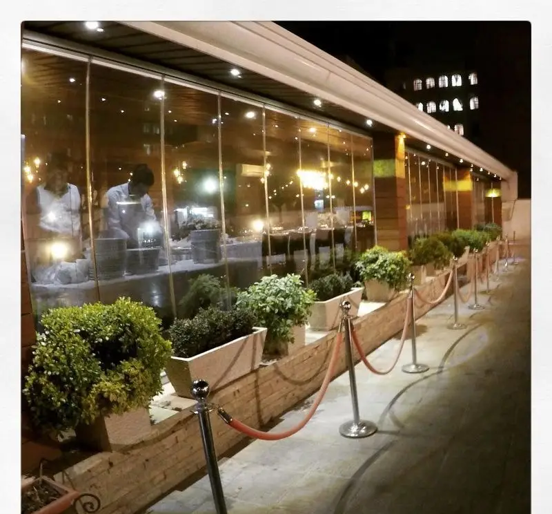 نمای بیرونی رستوران نایب ابان در شب با شیشه‌های نورانی، گیاهان گلدانی و طناب‌های قرمز لوکس. محیطی دلنشین برای صرف غذا.