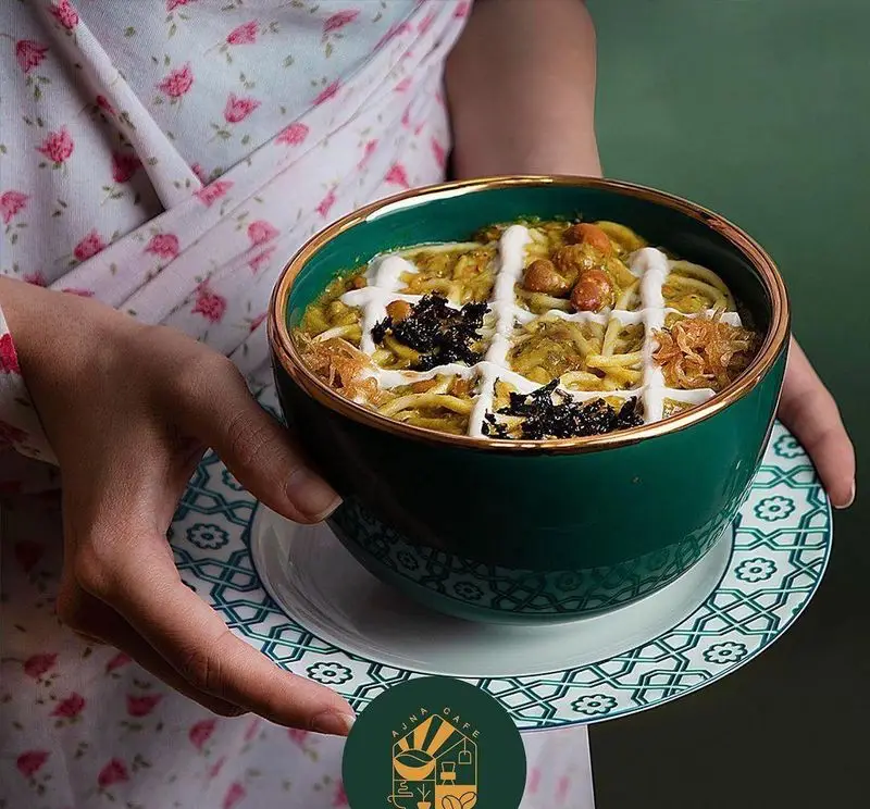 تصویری جذاب از آش رشته سنتی و خوشطعم کافه آجنا در کاسه زیبای سبز و طلایی با تزیین خاص کشک، نعناع و پیاز داغ.