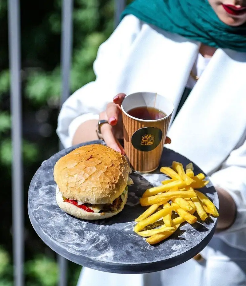 برگر تازه، سیب زمینی سرخ کرده طلایی و نوشیدنی گرم با لوگوی کافه آجنا در سینی مرمری شیک، آماده سرو برای میهمانان.