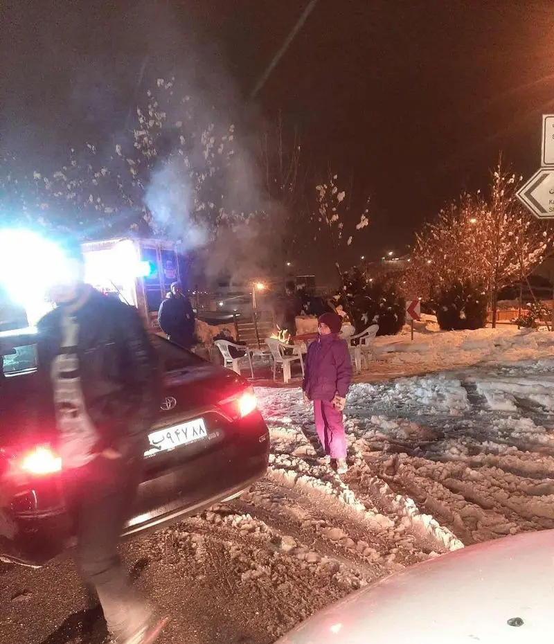 فضای باز کافه آجنا در شبی برفی، مشتریان در اطراف آتش گرمابخش دور هم جمع شده‌اند و از حال و هوای زمستان لذت می‌برند.