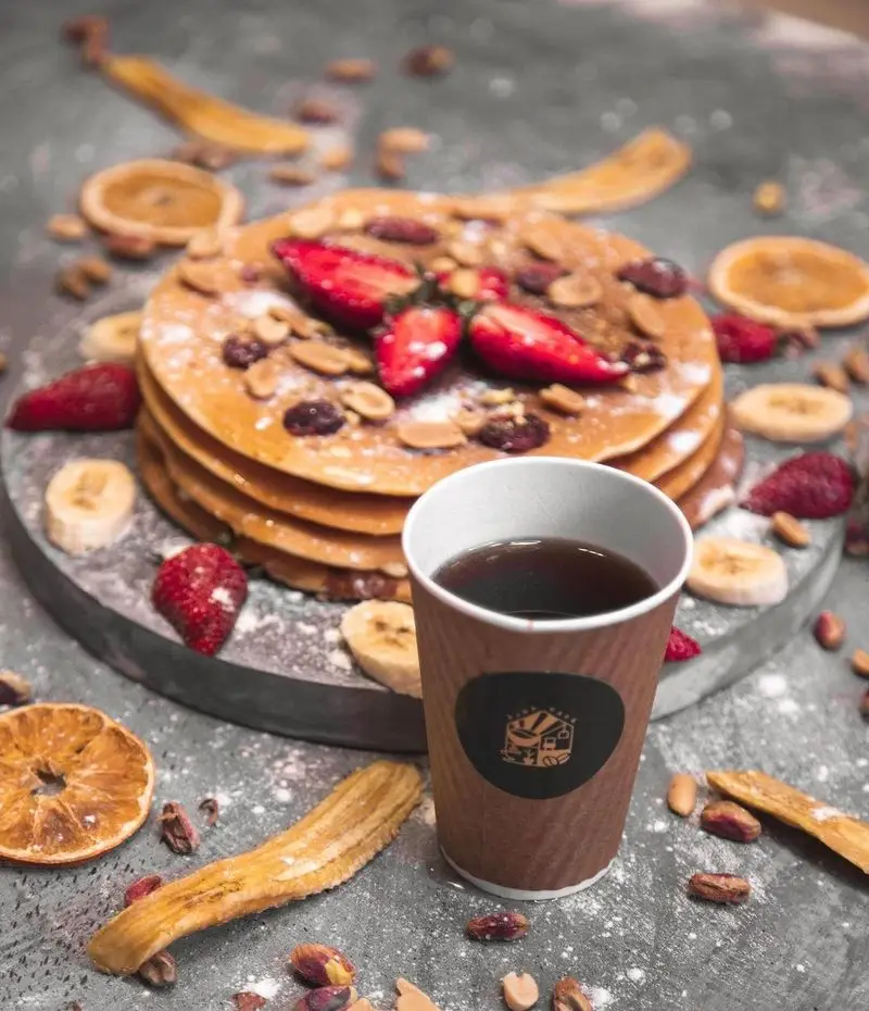 منوی صبحانه کافه آجنا: پنکیک‌های خانگی با توت فرنگی، موز خشک، پرتقال خشک، انواع آجیل و یک نوشیدنی گرم دلچسب.