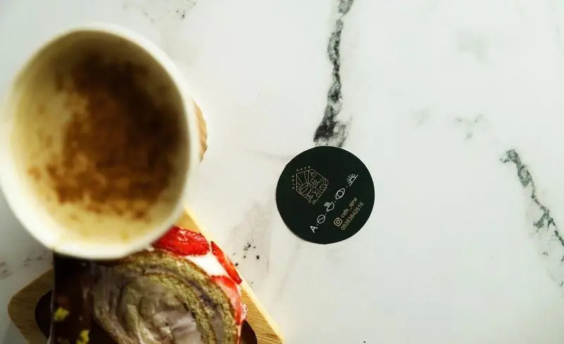 تصویر زیبا از یک فنجان نوشیدنی و رول کیک توت فرنگی خوشمزه روی میز مرمری، همراه با لوگو و شماره تماس کافه آجنا.