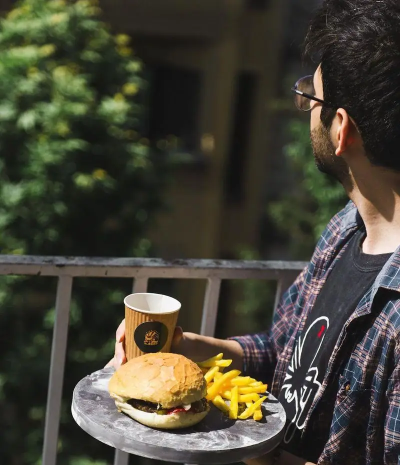 مشتری کافه آجنا در حال لذت بردن از یک وعده خوشمزه شامل همبرگر تازه، سیب زمینی سرخ کرده و نوشیدنی گرم در فضای باز و دلنشین بالکن.