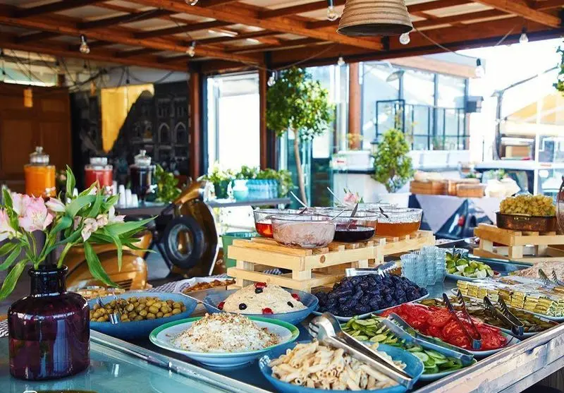 تصویری از میز بوفه گسترده کافه شمرون شامل انواع غذا، سالاد، میوه و دسرهای جذاب در محیطی دلپذیر و نورگیر.