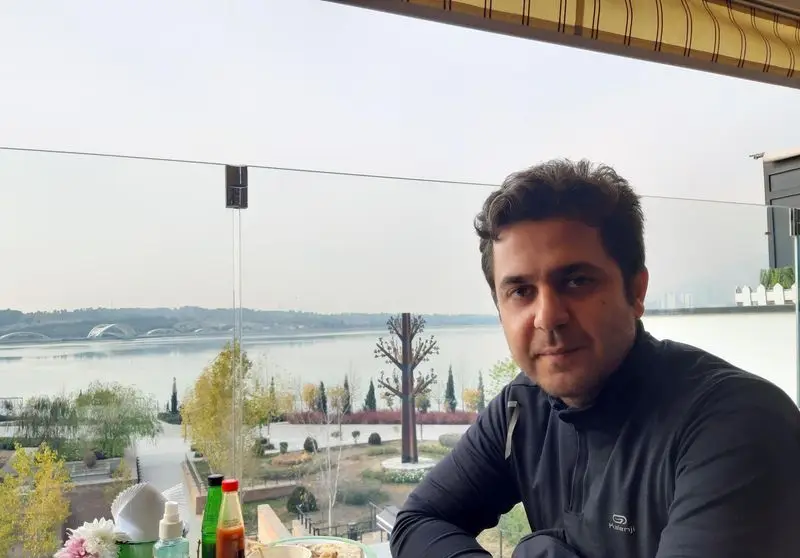 تصویری زیبا از کافه شمرون: مردی نشسته در بالکن کافه با منظره‌ای چشم‌نواز از دریاچه و فضای سبز اطراف، تجربه لذت‌بخش کافه‌گردی.