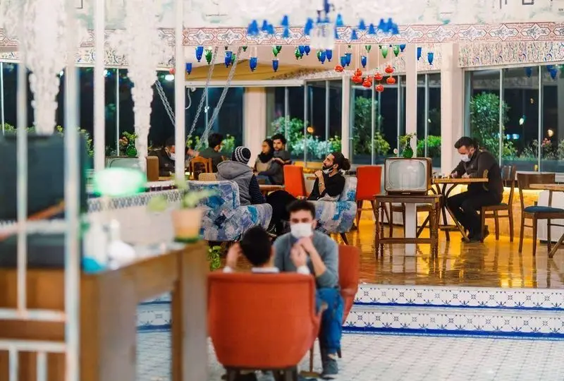 محیط داخلی کافه شمرون با مشتریانی در حال گپ و گفتگو، دکوراسیون دلنشین و چراغ‌های رنگی، فضایی دنج برای جمع‌های دوستانه.