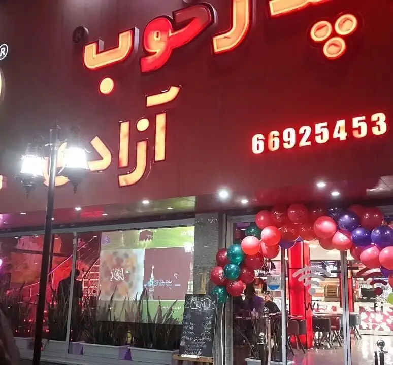 نمای بیرونی فست فود پدرخوب در شب با تابلوی نئون قرمز "پدرخوب آزاد" و شماره تماس 66925453. ورودی تزئین شده با بادکنک و تخته منو.