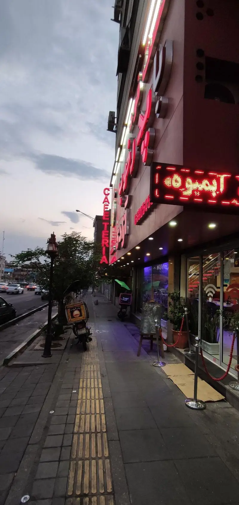 نمای بیرونی فست فود و کافه پدرخوب در یک خیابان شهری هنگام عصر، با تابلوهای نئون قرمز و LED. موتورسیکلت تحویل غذا در پیاده‌رو.