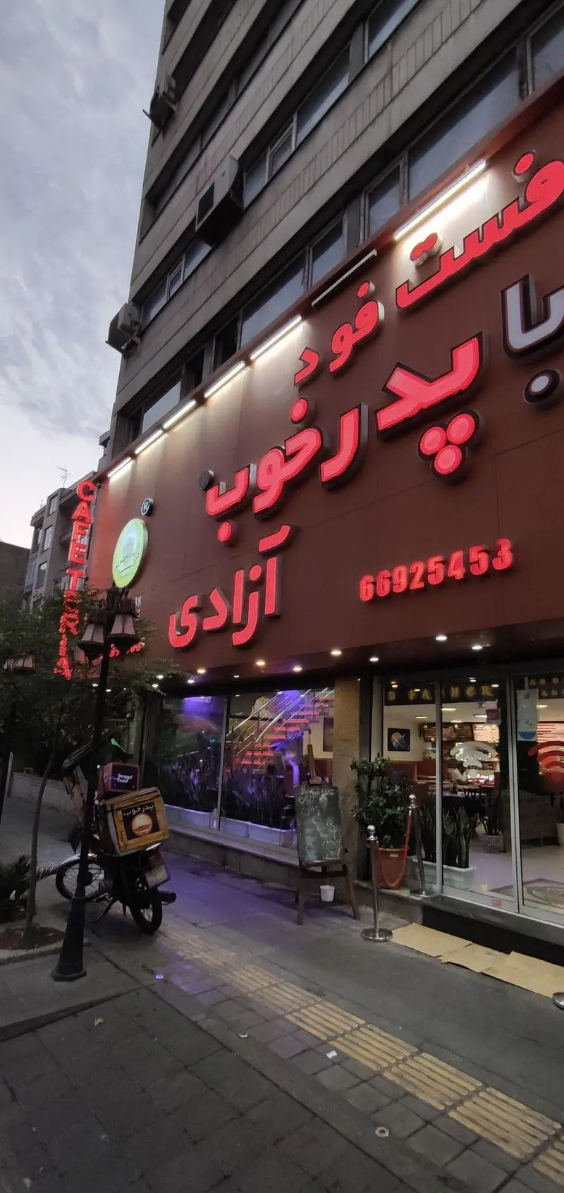 نمای بیرونی فست فود پدرخوب شعبه آزادی در تهران با تابلوی نئون قرمز روشن، شماره 66925453 و موتورسیکلت بیرون‌بر.