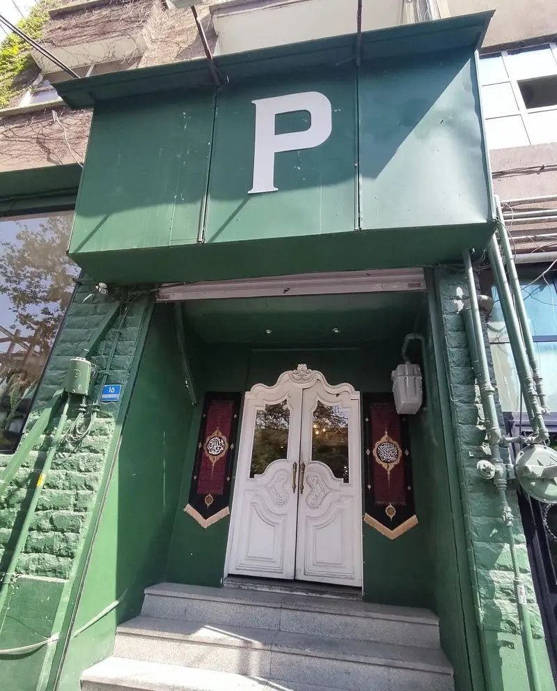 ورودی چشمگیر رستوران پیتزا پای با نمای سبز و تابلوی حرف P بزرگ سفید. درب سفید زیبا با تزئینات اسلامی و پله‌های سنگی.