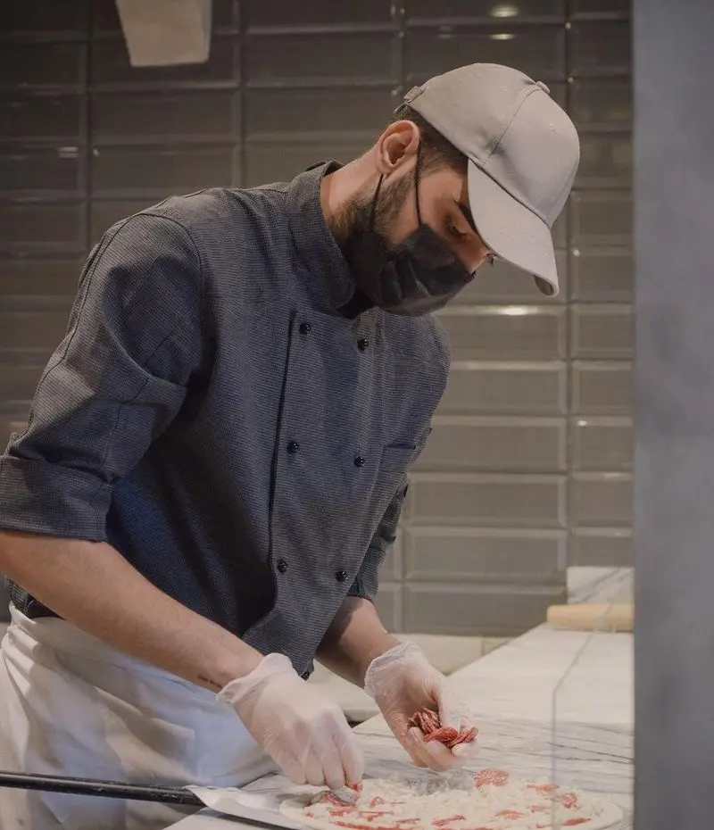 سرآشپز پیتزا پای در حال آماده‌سازی پیتزای تازه با مواد اولیه باکیفیت و رعایت کامل نکات بهداشتی (ماسک و دستکش).