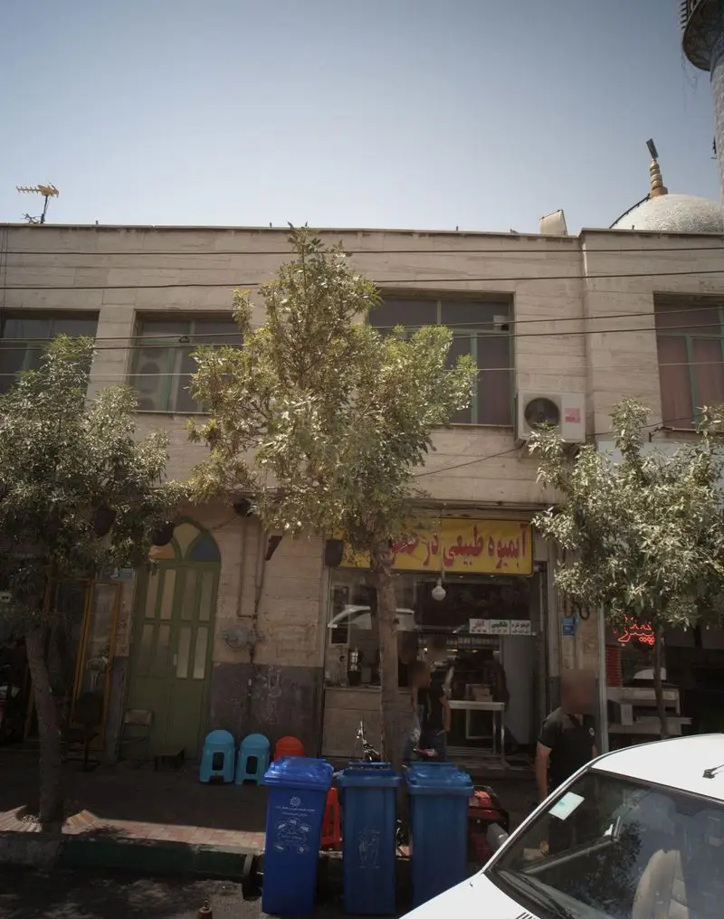 نمای بیرونی مغازه آبمیوه مهدی، عرضه کننده آبمیوه طبیعی، با تابلوی زرد رنگ و سطل‌های زباله آبی در یک خیابان شهری.