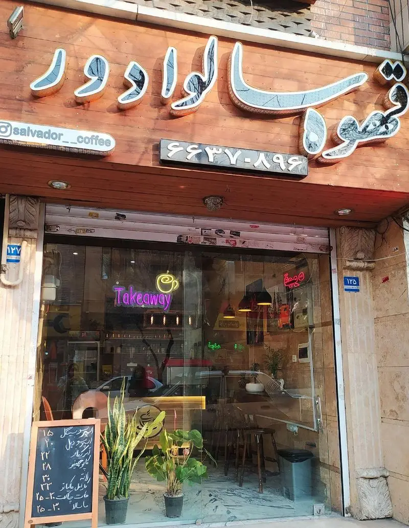 نمای بیرونی کافی شاپ قهوه سالوادور با تابلوی درخشان، شماره تماس و آدرس اینستاگرام @salvador_coffee. منوی نوشیدنی ها و تابلوی Takeaway در ویترین شیشه ای.