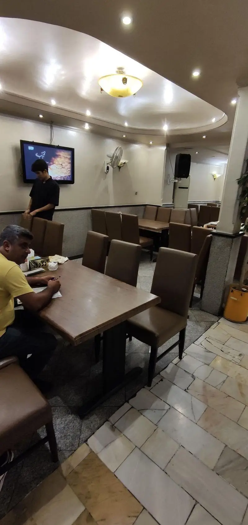 فضای داخلی چلوکبابی نصرت با میز و صندلی‌های چیده شده، مشتری در حال انتخاب غذا و کارکنان آماده خدمت. نورپردازی دلنشین و تلویزیون دیواری.