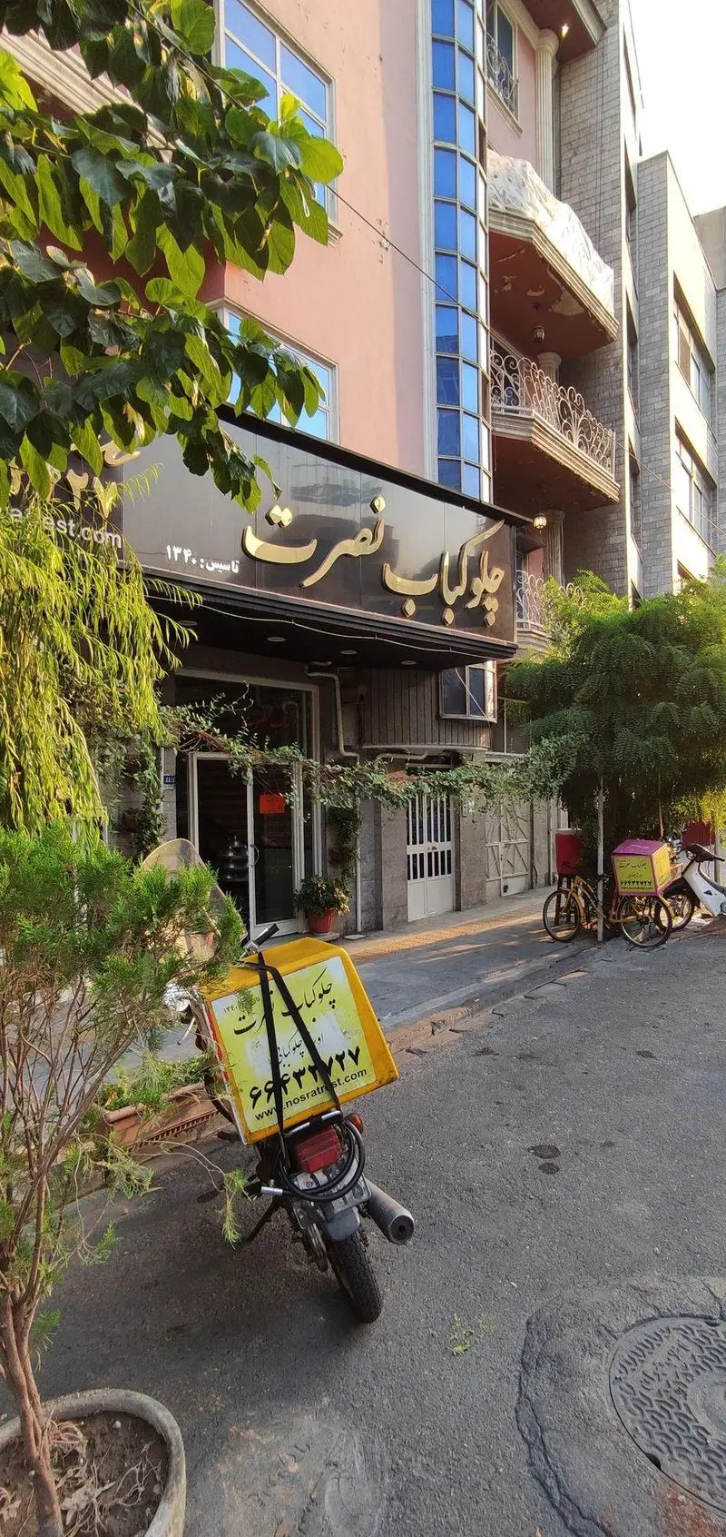 نمای بیرونی چلوکبابی نصرت با تابلوی طلایی، موتورسیکلت‌های پیک تحویل غذا و دوچرخه در کنار درختان سرسبز.