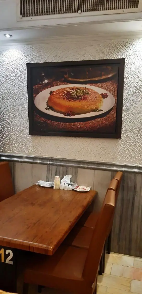 فضای داخلی گرم و صمیمی چلوکبابی نصرت، شامل میزهای چوبی، صندلی‌های قهوه‌ای و تابلوی یک ته چین زعفرانی دلپذیر ایرانی.