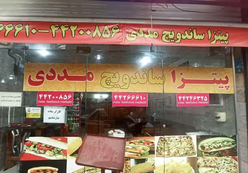 نمای بیرونی فست فود مددی، متخصص در پیتزا و ساندویچ. شامل تابلوهای تبلیغاتی، شماره تماس و اینستاگرام fastfood.madadiii.