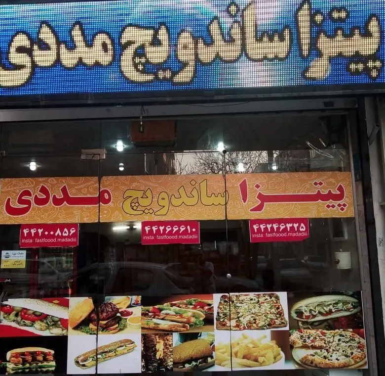 نمای بیرونی فست فود مددی با تابلوی روشن «پیتزا ساندویچ مددی»، شماره تماس‌ها و اینستاگرام. ویترین انواع پیتزا، ساندویچ، برگر و سیب‌زمینی سرخ‌کرده.