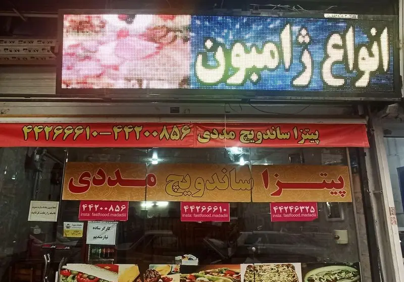 نمای بیرونی فست فود مددی: تابلوی LED نمایش‌دهنده انواع ژامبون، پیتزا، ساندویچ. شامل شماره تماس ۴۴۲۲۰۰۸۵۶ و آیدی اینستاگرام fastfoood.madadiii