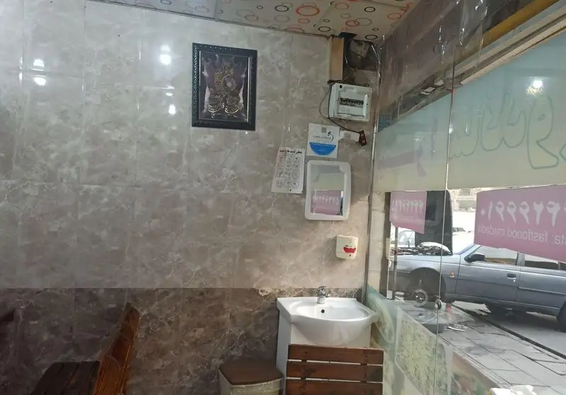 نمای داخلی فست فود مددی با دیوارهای کاشی‌کاری، سینک، و دکوراسیون ساده. بازتاب نام "فست فود مددی" روی شیشه و ماشین در بیرون.