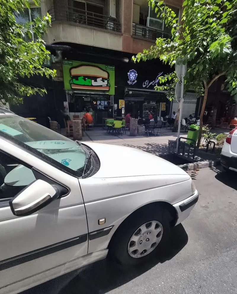 نمای بیرونی رستوران فست فود پک مک در یک روز آفتابی، با تابلوی بزرگ همبرگر، مشتریان در فضای باز و ماشین پارک شده.