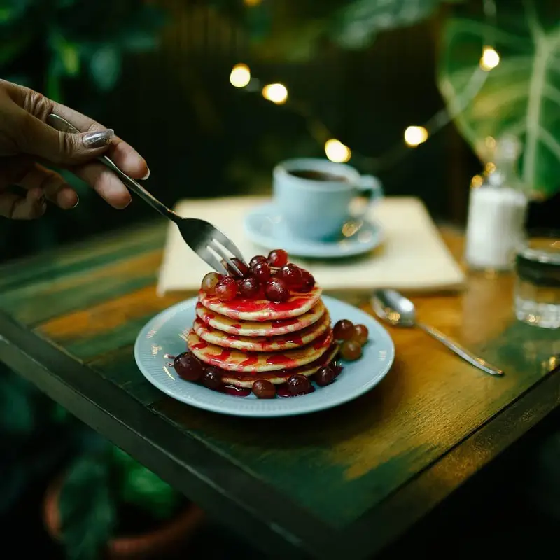 پنکیک‌های خوشمزه کافه ث با سس میوه و انگور روی میز چوبی، همراه قهوه داغ. ایده‌آل برای صبحانه‌ای دلپذیر و تجربه‌ای شیرین در کافه.